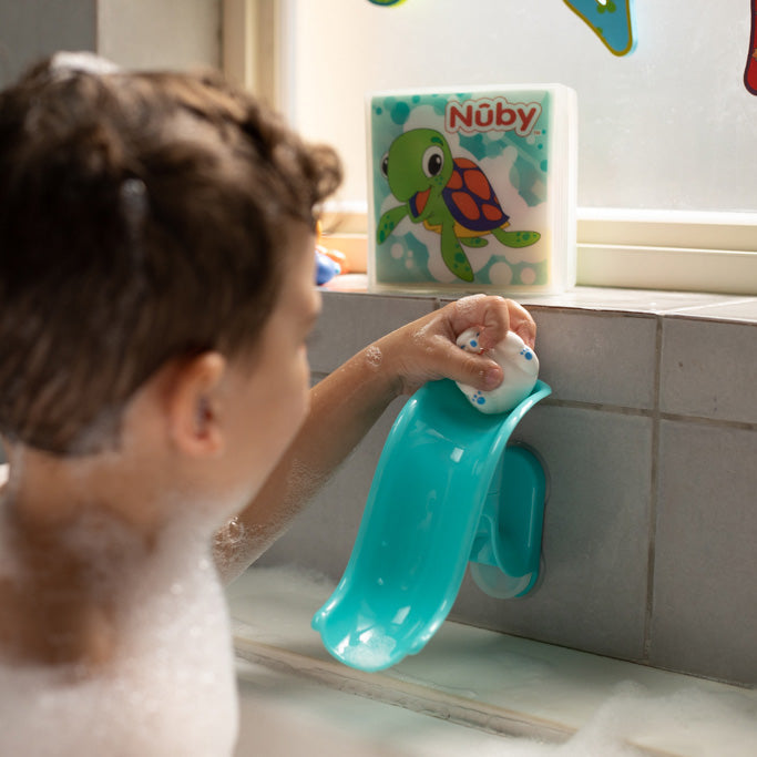 A child in a bubble bath plays with a Penguin Slide Squirt Bath Toy attached to the bath wall to boost hand-eye coordination, while a colorful Nuby turtle bath book sits on the windowsill in the background.