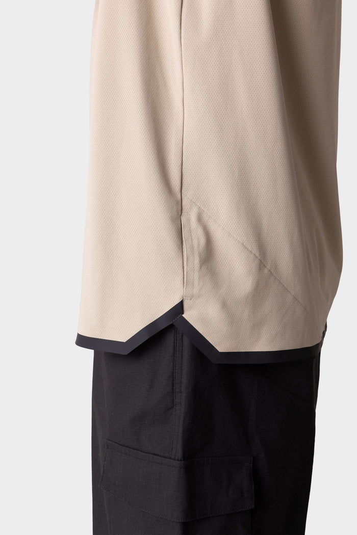 Close-up side view of a person wearing the 686 Mens Lets Go Tech Hoody in beige with a notched hem and black trim, paired with lightweight black cargo pants. The background is plain and light-colored. Brand: 686.