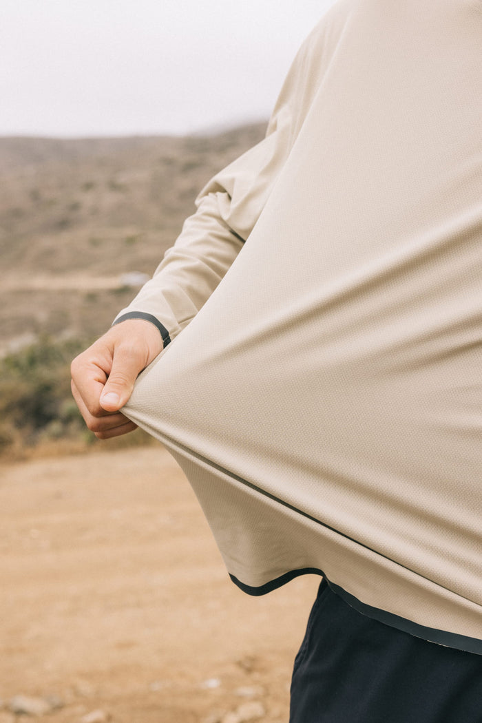 A person outdoors stretches the lightweight, beige 686 Mens Lets Go Tech Hoody away from their body. The background is a blurred, dry landscape with hills and scattered vegetation.
