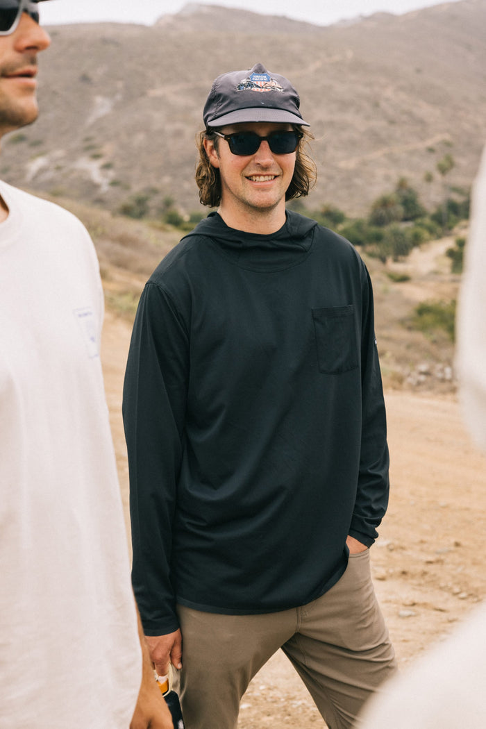 A man wearing sunglasses, a dark cap, and the 686 Mens Lets Go Tech Hoody stands outdoors on a dirt path with hills behind him. Other people are partially visible nearby.
