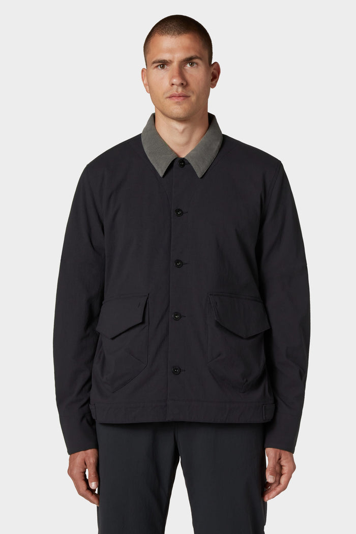 A man with short hair wears the 686 Men's Unwork® Everywhere Jacket by 686, featuring a black color, gray collar, and two front flap pockets, standing against a plain light background.