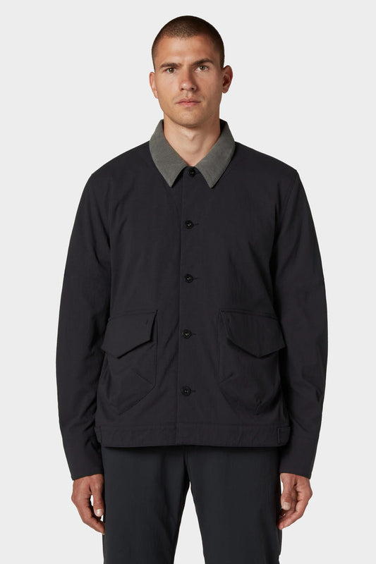 A man with short hair wears the 686 Men's Unwork® Everywhere Jacket by 686, featuring a black color, gray collar, and two front flap pockets, standing against a plain light background.