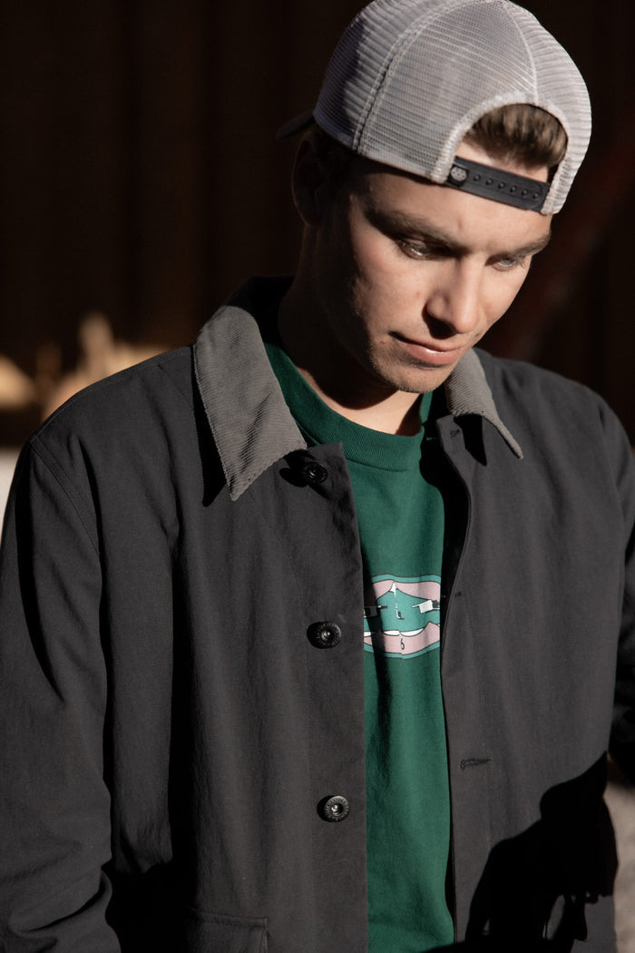 A young man in a backward white mesh cap, green graphic shirt, and the 686 Men's Unwork® Everywhere Jacket by 686 gazes down thoughtfully in gentle natural light.