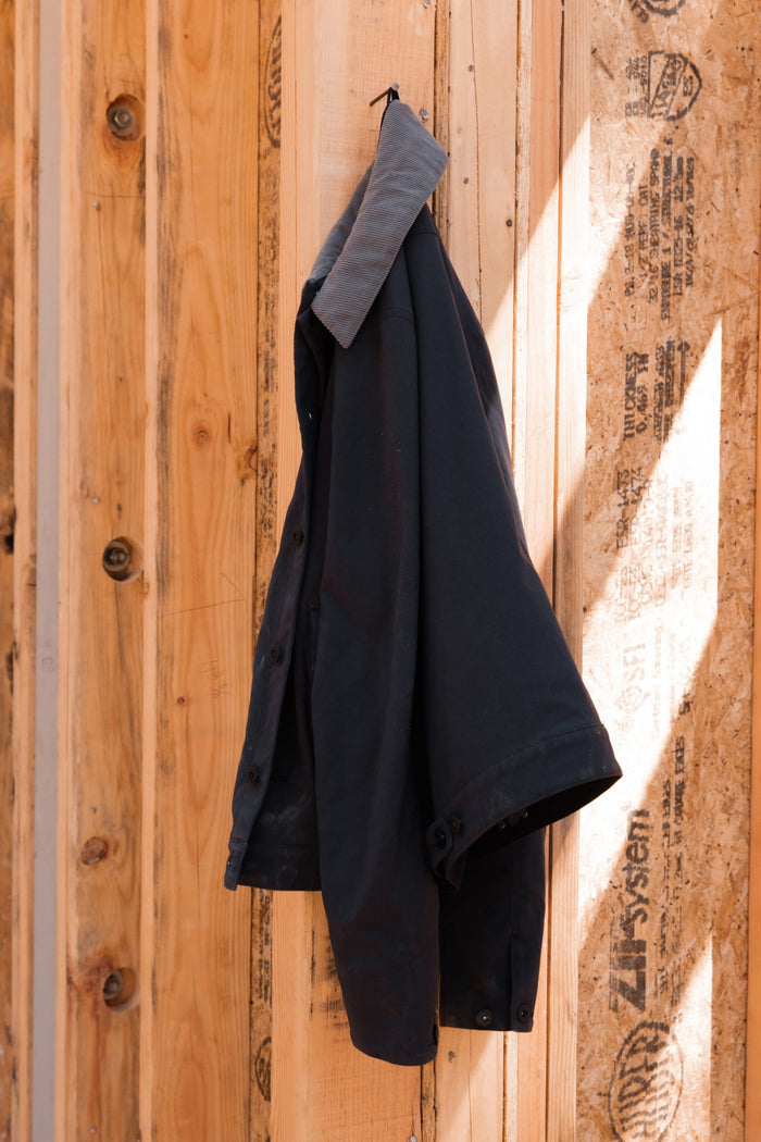 The 686 Men's Unwork® Everywhere Jacket by 686 hangs on a hook against a wall of unfinished wooden planks and oriented strand board, with sunlight casting intricate shadows across the natural wood backdrop.