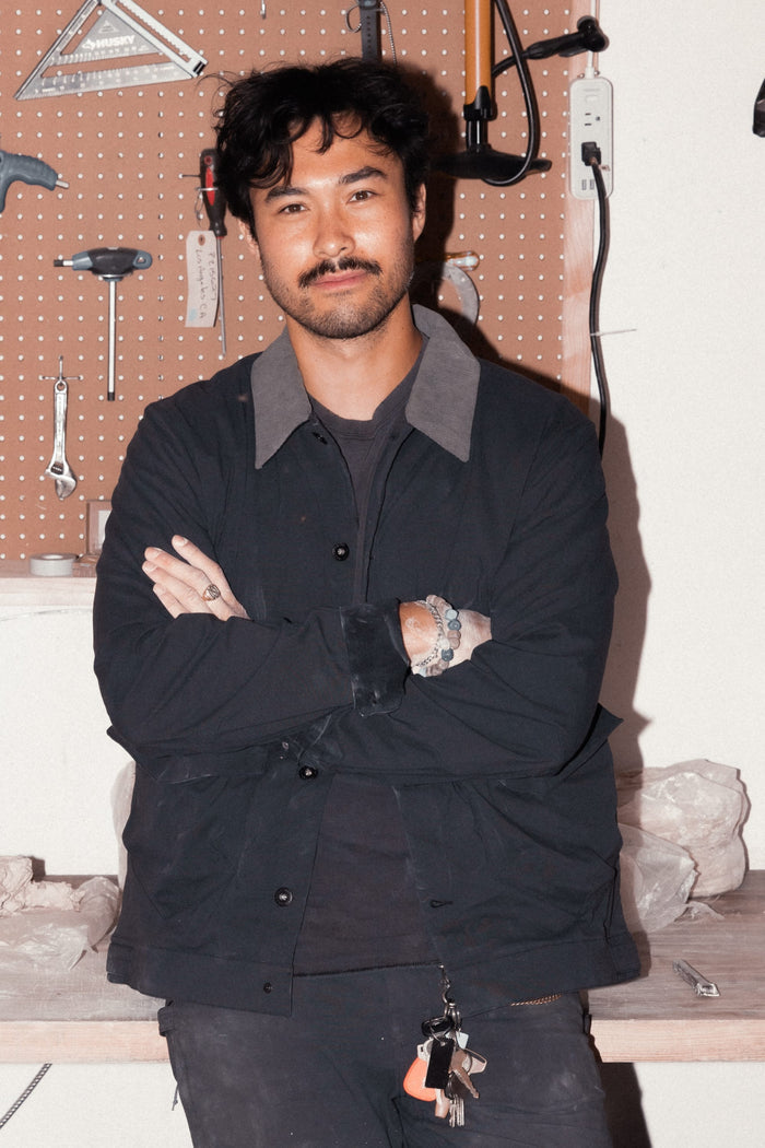 A person with dark hair and a mustache stands before a pegboard of tools, arms crossed, wearing the 686 Men's Unwork® Everywhere Jacket by 686 and a bracelet, with keys hanging from their pocket.