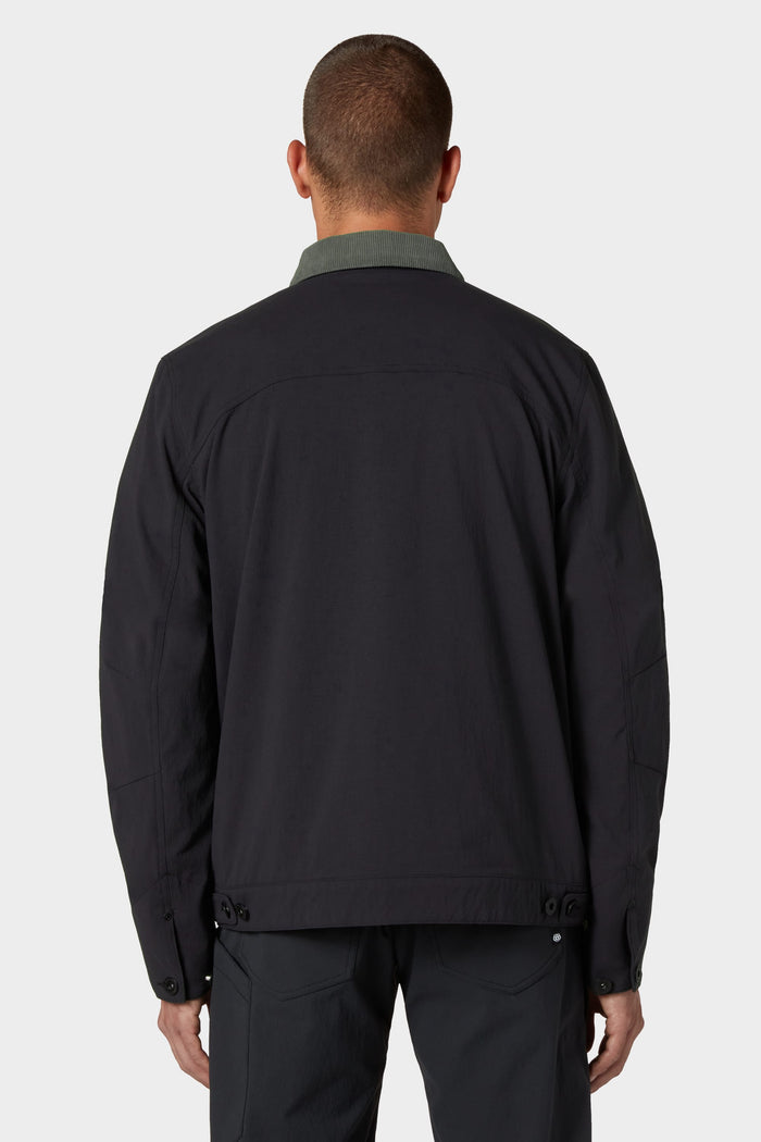 A person with short hair is seen from behind, wearing the 686 Men's Unwork® Everywhere Jacket by 686. The dark jacket features button details on the lower sides and a lighter collar. They are also wearing matching pants against a plain light gray background.