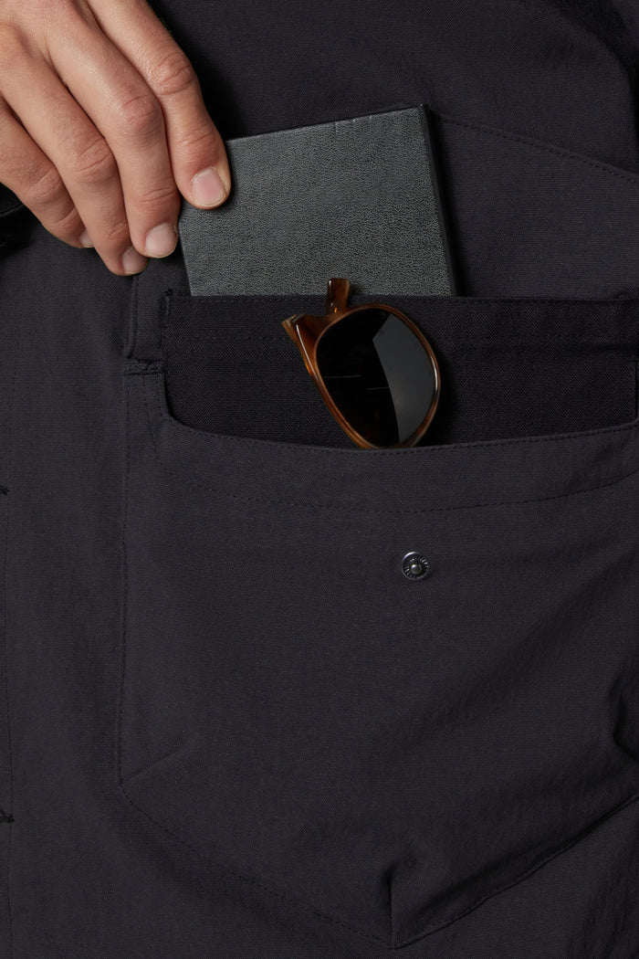 A hand places a closed black passport and brown sunglasses into the front pocket of the 686 Men's Unwork® Everywhere Jacket.