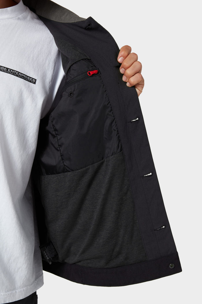 A person opens a black 686 Men's Unwork® Everywhere Jacket, showing its inner lining and large pocket. They're wearing a white T-shirt with partial chest text, set against a plain, light background.