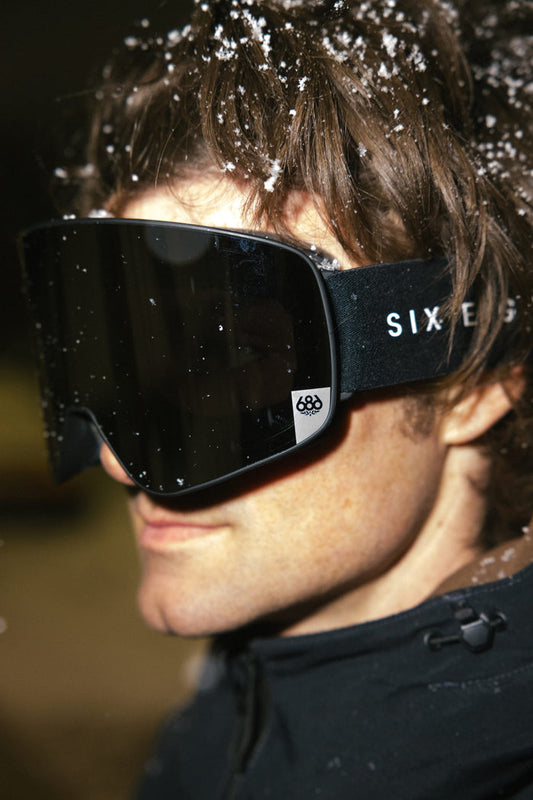 A person wearing the 686 SMARTY® Magnetic Goggle, with the 686 logo visible on the side, dressed in a dark jacket, snowflakes resting on their hair and goggles.