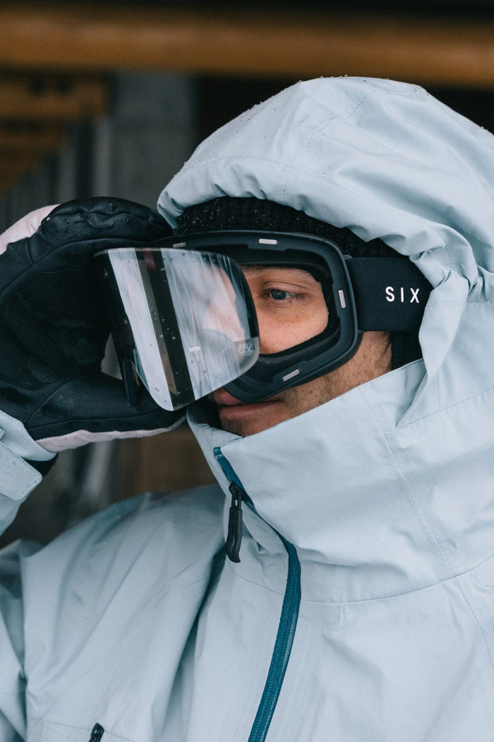 Wearing a light blue winter jacket and black gloves, a person adjusts their 686 SMARTY® Magnetic Goggle, with the word 686 visible on the side strap. Snow or frost is reflected on the goggles.