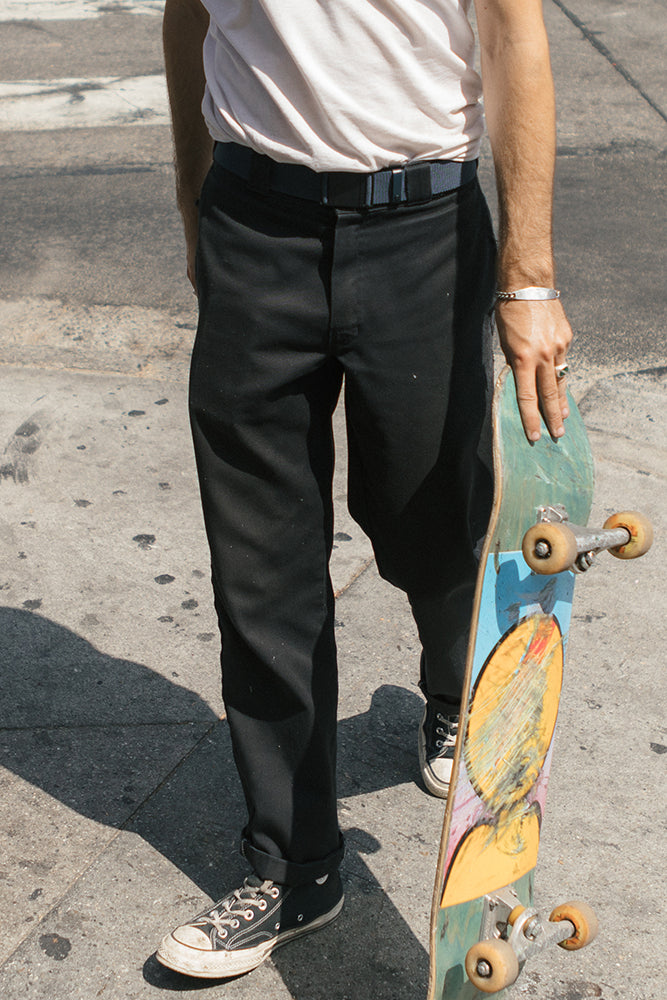A person wearing black pants, black Converse sneakers, a light t-shirt, and the 686 Westwell Clicker Stretch Belt stands on a sidewalk holding a skateboard with colorful artwork on its underside.