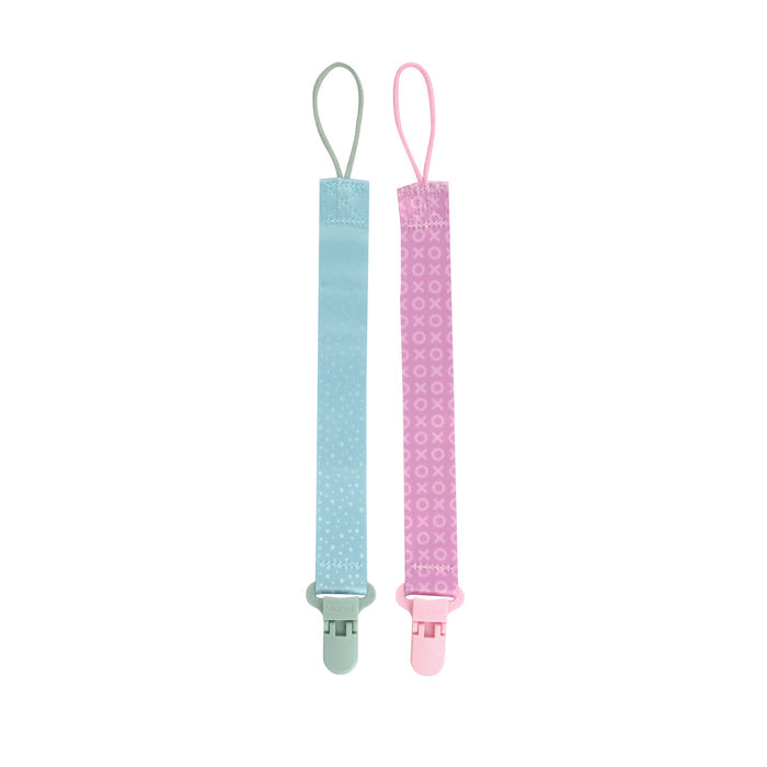 The Cloth Pacifier Clip (2 Pack) includes one light blue clip with white dots and one pink clip with X and O patterns, each featuring a top loop and bottom plastic clip for securing pacifiers. Shown on a white background.