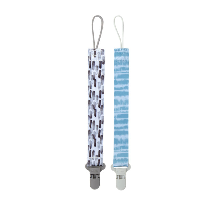The Cloth Pacifier Clip (2 Pack) includes two fabric-strap clips: one with a black and white geometric design and gray clip, the other with blue and white stripes and a white clip. They function as both pacifier holders and baby accessories.