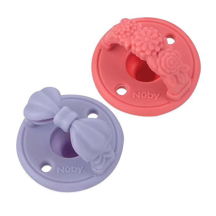 The Sili Soother Pacifier (2 Pack) includes two round silicone pacifier holders by Nuby—one pink with a floral design, one purple with a bow—each with three holes and a central opening, designed for Sili Soother Pacifiers.