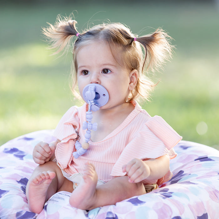 A baby with light brown pigtails sits on a floral cushion outdoors, wearing a pink ruffled top and holding the Silicone and Wood Beaded Pacifier Clip. The green background is softly blurred.