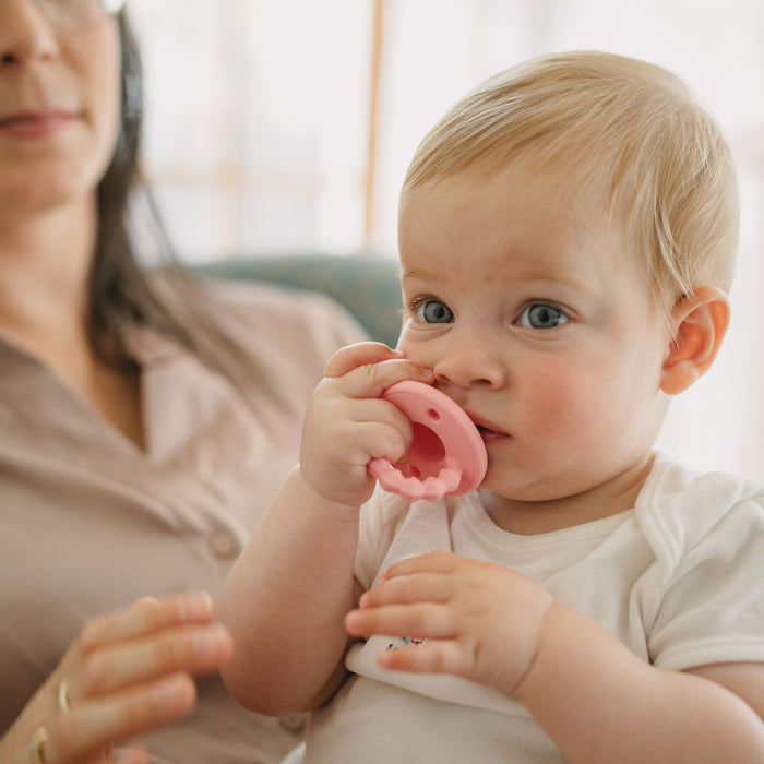 A blonde-haired baby sits on an adult’s lap indoors, holding and chewing on a Silicone Softees Pacifier & Teether (5 Pack). The adult in glasses and a beige shirt is partially visible under the soft natural light.