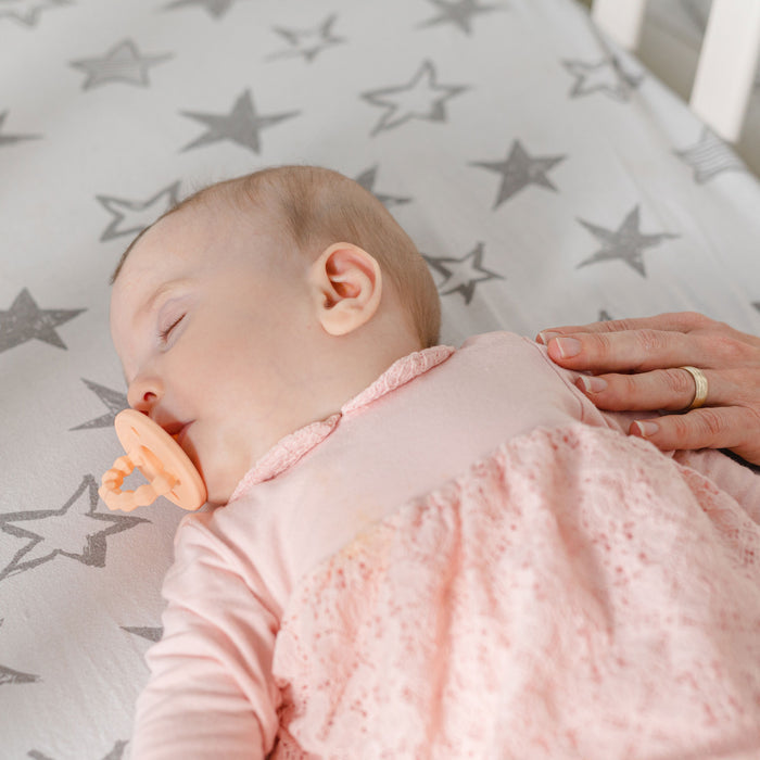 A baby in pink sleeps on a star-patterned sheet with a Silicone Softees Pacifier & Teether (5 Pack), while an adult hand gently rests on the baby's chest.