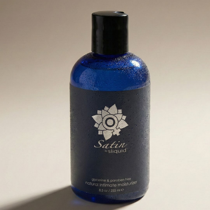 Angled close-up shot showing the water-droplet covered cobalt blue bottle of Sliquid Satin natural intimate moisturizer lying on a neutral surface.