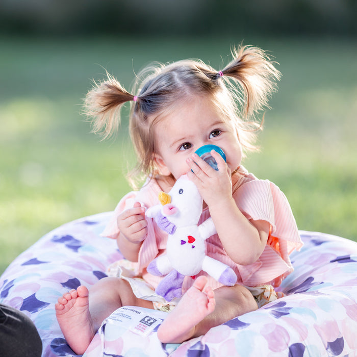 A toddler with pigtails sits outdoors on a patterned cushion, holding a white stuffed unicorn and soothing baby’s gums with the Snuggleez Pacifier and Clip Combo Set, with grass softly blurred in the background.