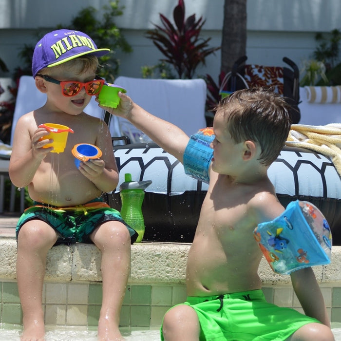 Two boys in swim trunks and floaties play by a pool with Splish Splash Stacking Cups - 5 pack, laughing as one pours water over the other’s head. The boy getting wet wears sunglasses and a purple cap; pool toys are visible in the background.