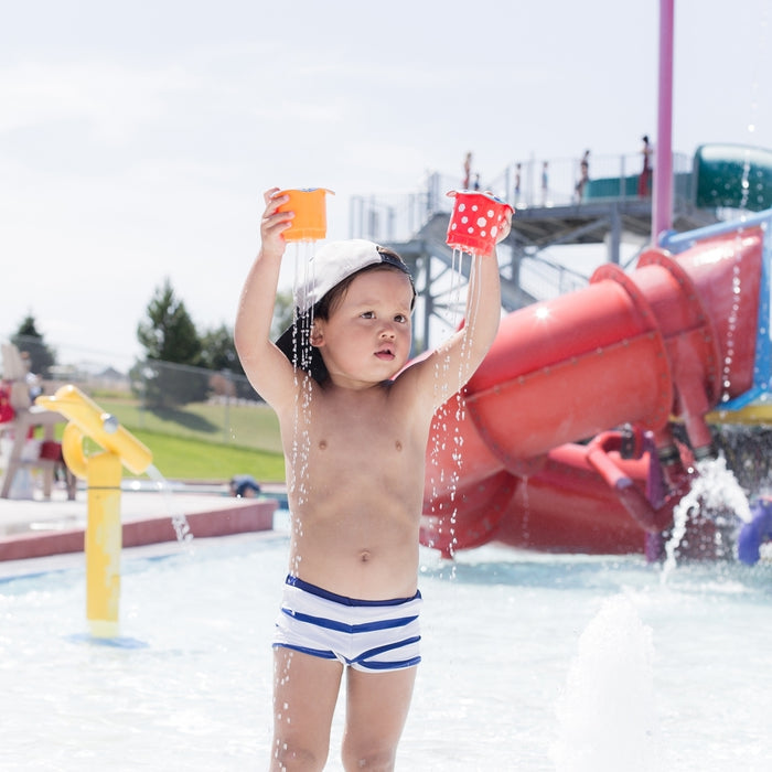A young child in striped swim trunks plays at a splash park, holding the Splish Splash Stacking Cups - 5 pack above their head as water pours out. Red slides and water features appear in the background.