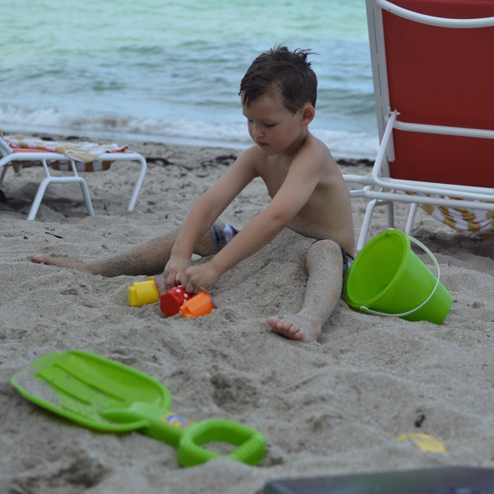 A young boy plays on the sandy beach with Splish Splash Stacking Cups - 5 pack. A green bucket and shovel are nearby, as ocean waves roll behind him and a red and white beach chair sits in the background.