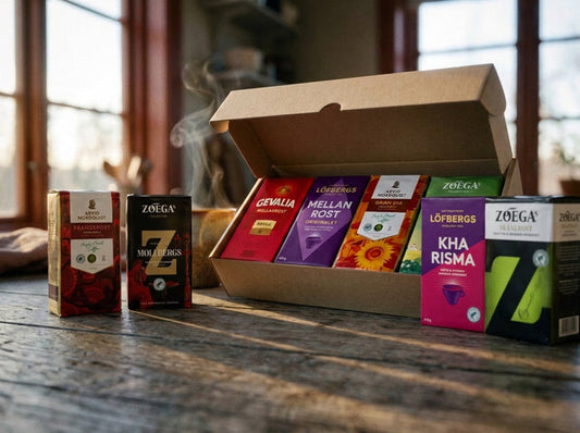 Swedish Coffee Taste Box Medium open on a rustic wooden table in warm morning light, showing 8 Swedish ground coffee bags – Gevalia Mellanrost, Löfbergs Mellanrost Originalet, Arvid Nordquist Gran Dia, Zoégas Fikastund, Zoégas Mollbergs Blandning, Löfbergs Kharisma, Zoégas Skånerost and Arvid Nordquist Franskrost – arranged in and around an open kraft box with a steaming mug in the background
