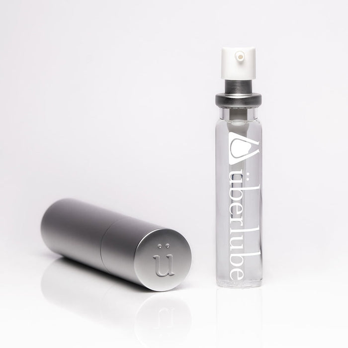 A tall, slender silver refillable case stands next to a clear glass bottle of "überlube" with a white pump top, both items for personal lubricant available at Worldcondoms.